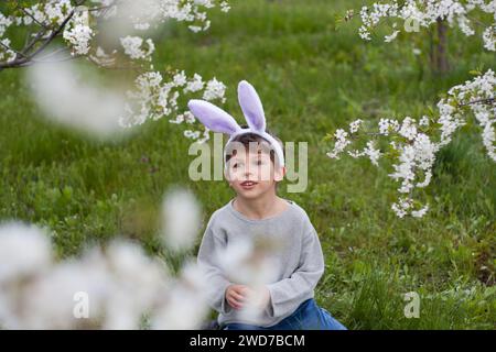 Mignon garçon préscolaire avec des oreilles de lapin dans le jardin. enfant souriant assis sur la pelouse verte sur fond d'arbres fleurissant avec des fleurs blanches. Joyeuses pâques Banque D'Images