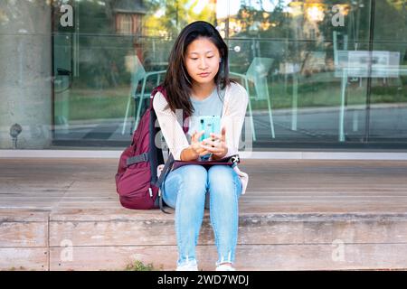 jeune femme asiatique assise naviguant sur internet sur un téléphone portable à l'extérieur. Banque D'Images