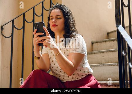 jeune femme latine avec des bigoudis, lisant attentivement les messages, les notifications des médias sociaux et les e-mails au téléphone, assis à l'extérieur dans les escaliers à la maison Banque D'Images