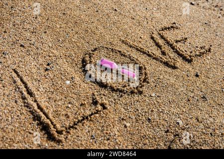 Amour intemporel : « l'amour » inscrit sur la plage, berçant un sablier rose, où la romance rencontre le sable des moments éternels Banque D'Images