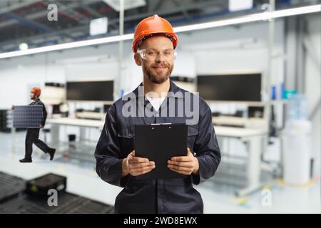 Technicien en uniforme avec un casque et des lunettes tenant une presse-papiers à l'intérieur d'une usine Banque D'Images