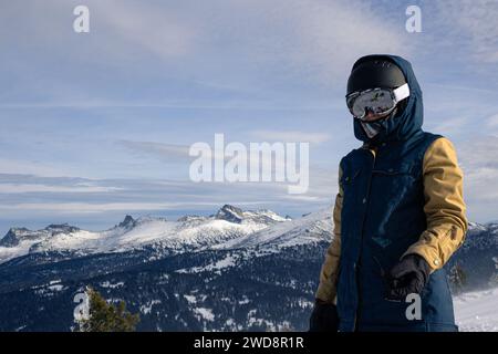 Femme dans l'équipement de ski ou de snowboard avec en toile de fond des montagnes et des sommets rocheux enneigés. Veste chaude avec membrane, gants, lunettes de ski blanches Banque D'Images