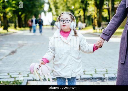 Enfant ayant besoin spécial tenant les mains de la mère portant des écouteurs s'amusant dans le parc de la ville en automne. Banque D'Images