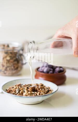 Main de femme versant le lait de la bouteille dans un bol de granola. Situé sur un fond clair avec un pot de granola et de myrtilles en arrière-plan. Banque D'Images
