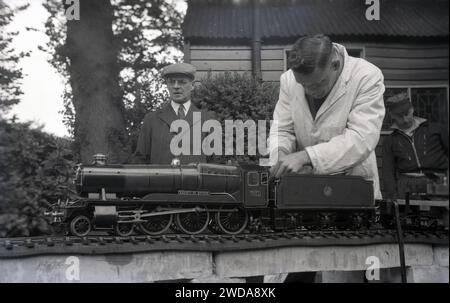 Années 1950, historique, à l'extérieur dans le jardin de la Field End Railway Co, Angleterre, Royaume-Uni, amateur de modèle de chemin de fer à vapeur en blouse blanche travaillant sur un train à vapeur miniature, «County of Oxford» (1023) une locomotive à vapeur GWR qui est entrée en service en 1947. La locomotive à vapeur County Class ou 1000 Class était le dernier développement de la classe Saint à deux cylindres introduite à l'origine en 1901. Banque D'Images