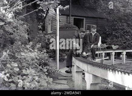 Années 1950, historique, à l'extérieur dans un jardin, amateurs de chemin de fer miniature dans un chemin de fer à vapeur de jardin miniature, à la Field End Railway Co, Angleterre, Royaume-Uni. Sur une voie surélevée assise sur des poteaux en béton, deux hommes montent sur de minuscules trains à vapeur, l'un étant un modèle réduit de la locomotive à vapeur GWR 'County of Oxford' (1023), dont l'original a commencé à fonctionner en 1947. Banque D'Images