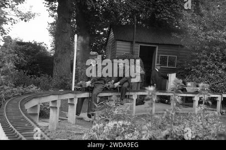 Années 1950, historique, à l'extérieur dans un jardin, amateurs de chemin de fer miniature dans un chemin de fer à vapeur de jardin miniature, à la Field End Railway Co, Angleterre, Royaume-Uni. Sur une voie surélevée assise sur des poteaux en béton, deux hommes montent sur de minuscules trains à vapeur, l'un étant un modèle réduit de la locomotive à vapeur GWR 'County of Oxford' (1023), dont l'original a commencé à fonctionner en 1947. Banque D'Images