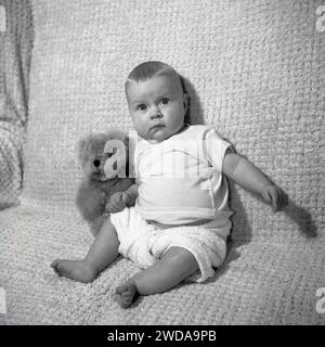 années 1960, historique, un petit garçon avec son ours en peluche à côté de lui assis sur une chaise recouverte de coton regardant la caméra. Banque D'Images