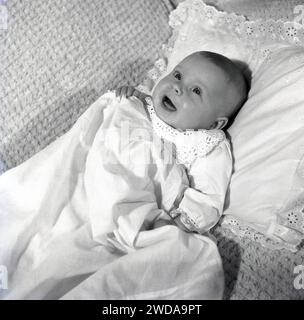 années 1960, historique, vue d'en haut d'un bébé garçon dans une tenue de baptême en dentelle, bouche et yeux grande ouverte tête allongée sur un oreiller sur une chaise recouverte de coton. Banque D'Images