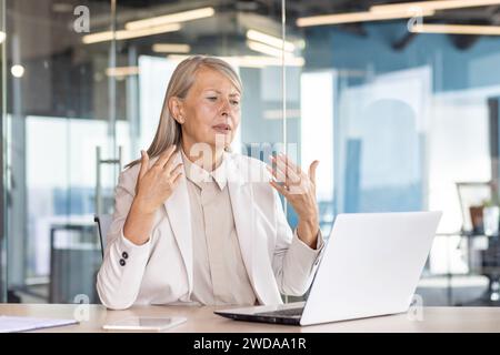 Femme senior sérieuse travaille dans le bureau, s'assoit à une table avec un ordinateur portable et agite ses mains dans son visage dans l'air de GET, il se sent mal. Banque D'Images