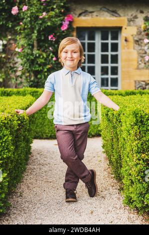 Adorable petit garçon posant dans le jardin classique anglais tapiary, portant un pantalon marron et une chemise de style vintage. Banque D'Images