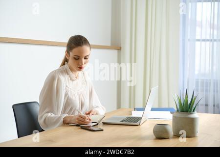 Concentré jeune femme en blouse blanche prenant des notes tout en travaillant au bureau avec ordinateur portable Banque D'Images
