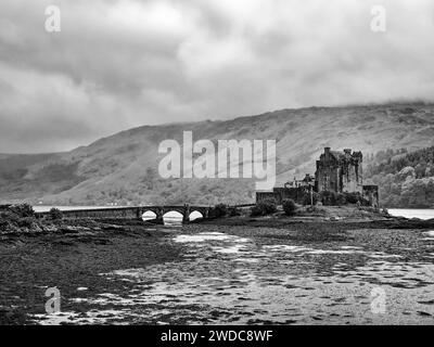 Photo en noir et blanc d'un vieux château avec pont devant un paysage montagneux et ciel nuageux, château d'Eilean Donan. Écosse, Grande-Bretagne Banque D'Images