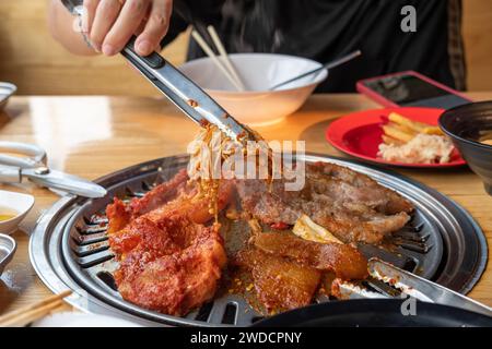 La main d'une femme cueille des champignons aux aiguilles dorées marinés en sauce sur un wok coréen sur un grill dans un restaurant. Banque D'Images