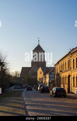Une vue paisible en soirée sur une rue pavée de Treuenbrietzen, avec des bâtiments historiques et une église sous le coucher de soleil doré. Banque D'Images