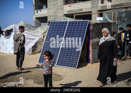 Rafah, Territoires palestiniens. 20 janvier 2024. Les Palestiniens marchent devant un panneau solaire, utilisé par certains pour produire de l’électricité dans les camps de réfugiés de Rafah. En raison de la détérioration de la situation financière des personnes et du manque d ' électricité dans les camps de réfugiés de Rafah, certains jeunes Palestiniens qui ont des panneaux solaires les utilisent pour gérer de petites entreprises grâce auxquelles ils permettent aux gens de recharger complètement leur téléphone pour environ 2 shekels israéliens (environ 55 cents US). Crédit : Mohammed Talatene/dpa/Alamy Live News Banque D'Images