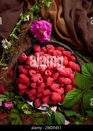 Framboises fraîches sur une assiette noire. Nature morte d'été avec, framboises, fleurs et herbes de prairie. Vue du dessus. Banque D'Images