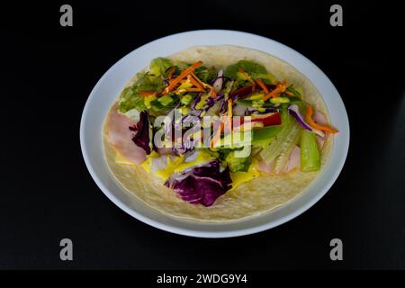 Jambon, mozzarella, laitue et poivrons rouges et verts tranches et croustilles de carottes sur tortilla de farine recouverte de moutarde au miel sur fond noir Banque D'Images