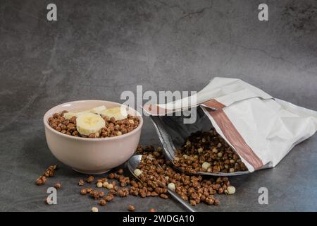 Bol de céréales aromatisées au chocolat croquant et bananes à côté du sac ouvert et cuillère sur fond texturé noir Banque D'Images