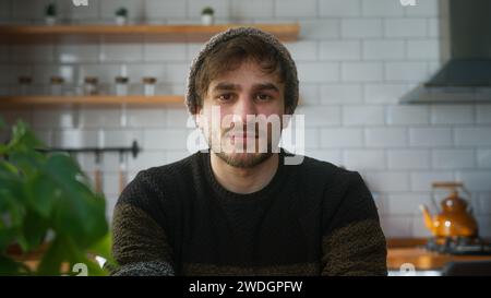 Portrait de jeune homme avec bonnet assis dans la cuisine moderne à la maison regardant la caméra ayant une conférence téléphonique, écoutant l'orateur sur la vidéo Banque D'Images