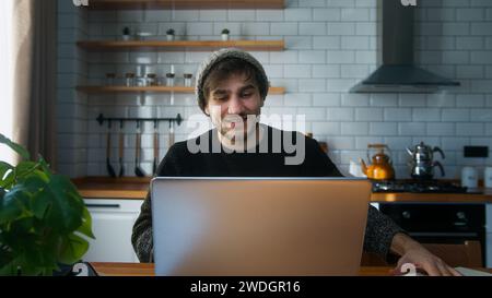 Jeune homme freelance adulte avec bonnet assis dans la cuisine moderne à la maison faire de la recherche, travailler avec un ordinateur portable et prendre des notes dans un cahier. Wor Banque D'Images