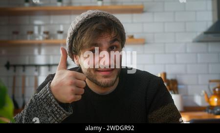 Portrait de jeune homme avec bonnet assis dans la cuisine moderne à la maison regarde la caméra et fait un signe pouce vers le haut Banque D'Images