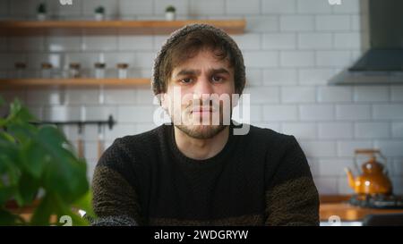 Un homme avec bonnet assis dans la cuisine moderne à la maison regardant la caméra ayant une conférence téléphonique, écoutant l'orateur sur l'appel vidéo. Webcam vie Banque D'Images