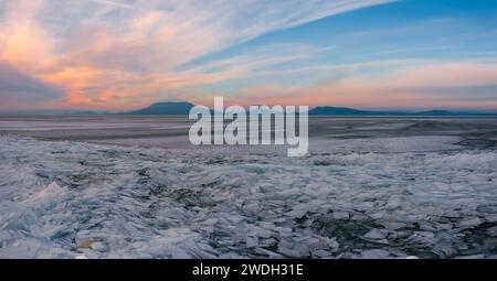 Fonyod, Hongrie - vue aérienne panoramique sur le lac gelé Balaton avec Badacsony et Gulacs en arrière-plan. Magnifique coucher de soleil nuageux d'hiver. Banque D'Images