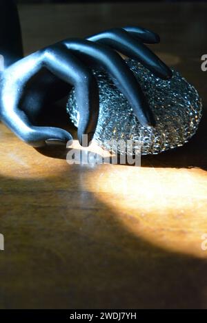 Statuette noire d'une main féminine mate avec une éponge métallique sur une table en bois brun de style ancien avec les rayons du soleil. Banque D'Images