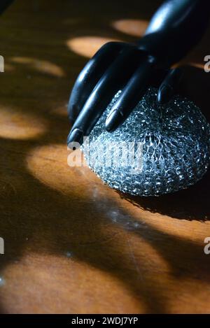 Statuette noire d'une main féminine mate avec une éponge métallique sur une table en bois brun de style ancien avec les rayons du soleil. Banque D'Images