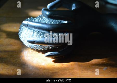 Statuette noire d'une main féminine mate avec une éponge métallique sur une table en bois brun de style ancien avec les rayons du soleil. Banque D'Images