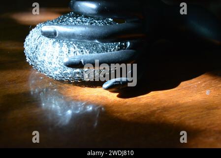 Statuette noire d'une main féminine mate avec une éponge métallique sur une table en bois brun de style ancien avec les rayons du soleil. Banque D'Images