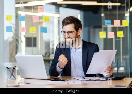 Homme d'affaires comptable prospère en costume avec des documents, des papiers, des rapports dans les mains travaille à l'intérieur du bureau au lieu de travail, financier masculin satisfait de la réalisation des résultats célèbre la victoire utilise un ordinateur portable. Banque D'Images