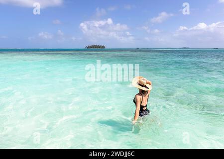 Femme touriste avec un chapeau de paille marchant en maillot de bain dans une eau de shalllow turquoise avec une île tropicale à l'arrière, San blas, mer des Caraïbes, Panama Banque D'Images