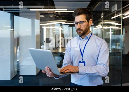 Homme d'affaires sérieux, administrateur système se tient avec un ordinateur portable dans ses mains, un homme pensant dans une chemise lit les données judicieusement, près de la fenêtre, un programmeur travaille à l'intérieur du bureau avec un ordinateur. Banque D'Images