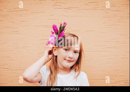 Portrait candide d'adorable petite fille de 6-7 ans tenant une fleur rose à côté de la tête Banque D'Images