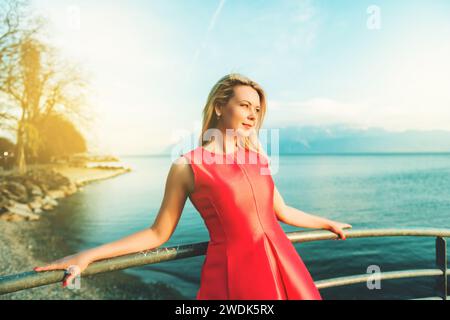 Portrait d'une jeune femme blonde posant à l'extérieur près du lac, vêtue d'une robe rouge vif Banque D'Images