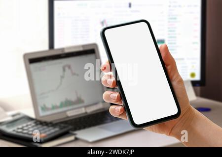 Téléphone intelligent avec écran vide en main devant les ordinateurs avec des graphiques financiers sur les moniteurs. Banque D'Images
