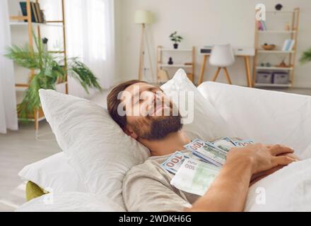 Homme endormi souriant couché dans son lit avec les yeux fermés, embrassant beaucoup de billets de banque en dollars à la maison Banque D'Images