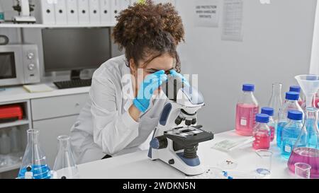 Femme hispanique avec les cheveux bouclés à l'aide d'un microscope dans un environnement de laboratoire, entourée d'équipement scientifique. Banque D'Images