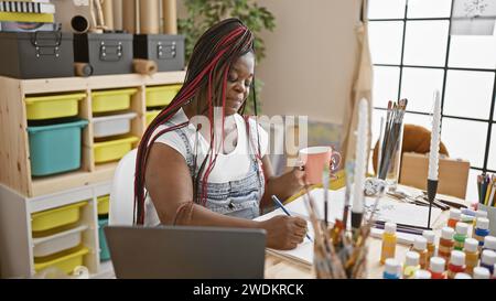 Belle artiste femme afro-américaine peignant un portrait, dessinant sur un cahier tout en buvant délicieusement le café du matin dans son studio d'art confortable Banque D'Images