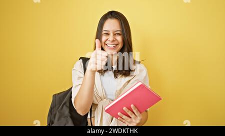 Portrait lumineux et joyeux d'une jeune, belle femme hispanique, clignotant avec confiance un geste de pouce vers le haut tout en étant absorbé dans la lecture de son livre scolaire, isol Banque D'Images