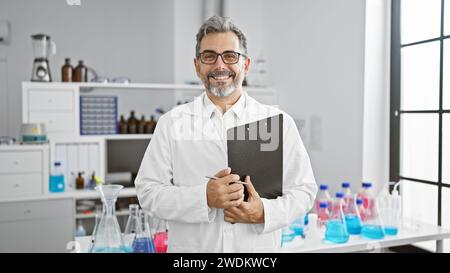 Jeune scientifique hispanique aux cheveux gris souriant confiant tenant le presse-papiers au laboratoire Banque D'Images