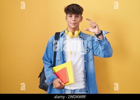 Adolescent hispanique portant un sac à dos étudiant et tenant des livres souriants et confiants gestuant avec la main faisant signe de petite taille avec les doigts regardant et Banque D'Images