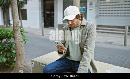 Jeune homme barbu dans des vêtements décontractés à l'aide d'un smartphone dans la rue de la ville Banque D'Images