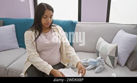 Jeune femme enceinte assise confortablement sur le canapé à la maison, se relaxant tout en pliant des vêtements de bébé Banque D'Images