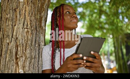 Femme afro-américaine confiante, décontracté élégant dans le parc de la ville, rayonne de positivité tout en utilisant le touchpad, son sourire prêtant un air de chee infectieux Banque D'Images