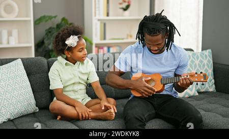 Père et fille afro-américains assis sur le canapé enseignant à jouer ukulele à la maison Banque D'Images