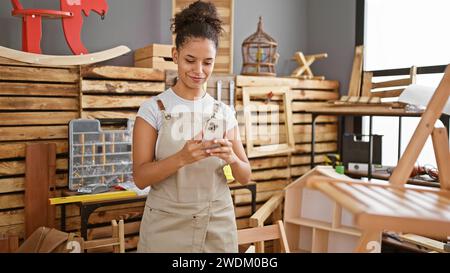 Souriante jeune femme hispanique charpentière, belle comme une image, capture en toute confiance son chef-d'œuvre de chaise en bois dans un atelier de menuiserie dynamique Banque D'Images