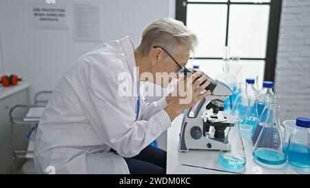 Scientifique senior dédiée aux cheveux gris immergée dans un travail intense de recherche au microscope au niveau du doctorat en laboratoire Banque D'Images